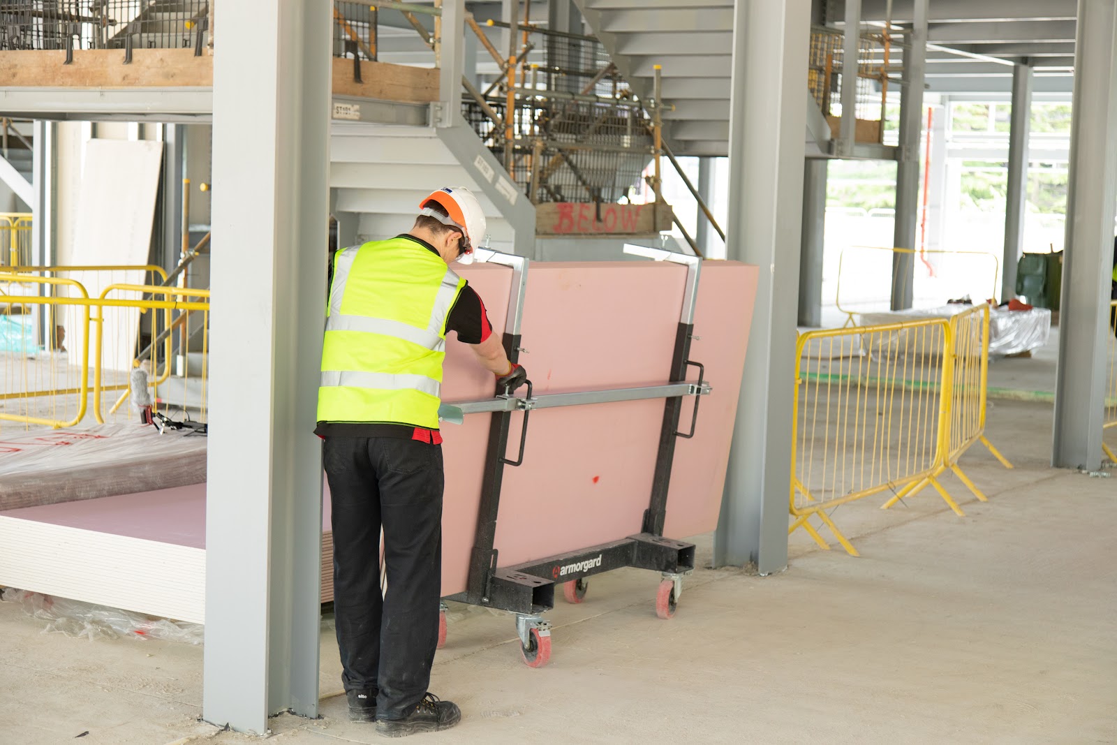 A person at a building site in a hi vis vest and hard had pulling a LoadAll trolly which has some sheet materials on it