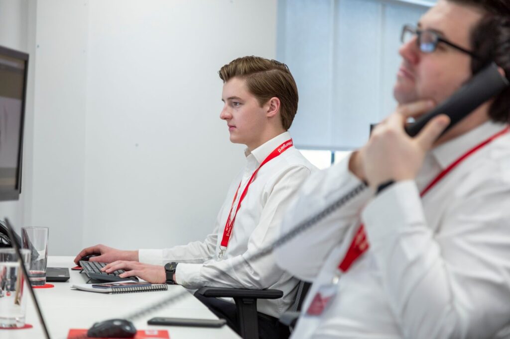 two people in an office setting, one person is looking at a computer screen and typing on the keyboard and the person next to them is on the phone