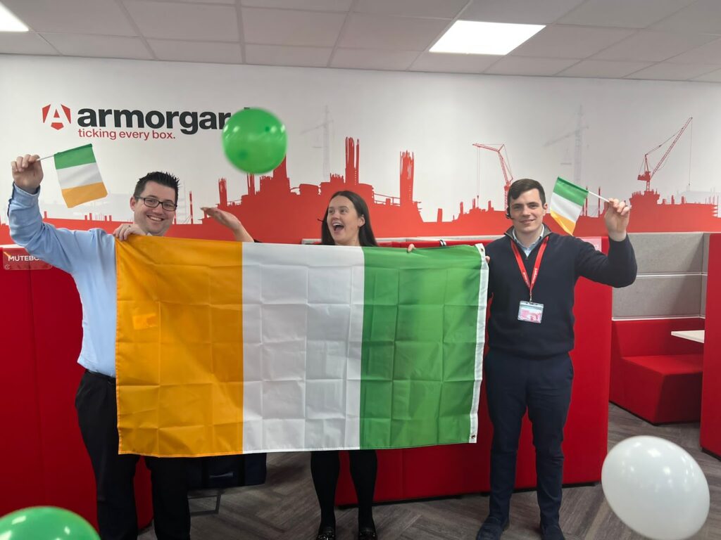 3 armorgard employees in the office person in the middle looks like they just thrown a green balloon in the air whilst holding a medium sized Irish flag, the two people on either end are holding the medium sized flag as well as smaller waving flags