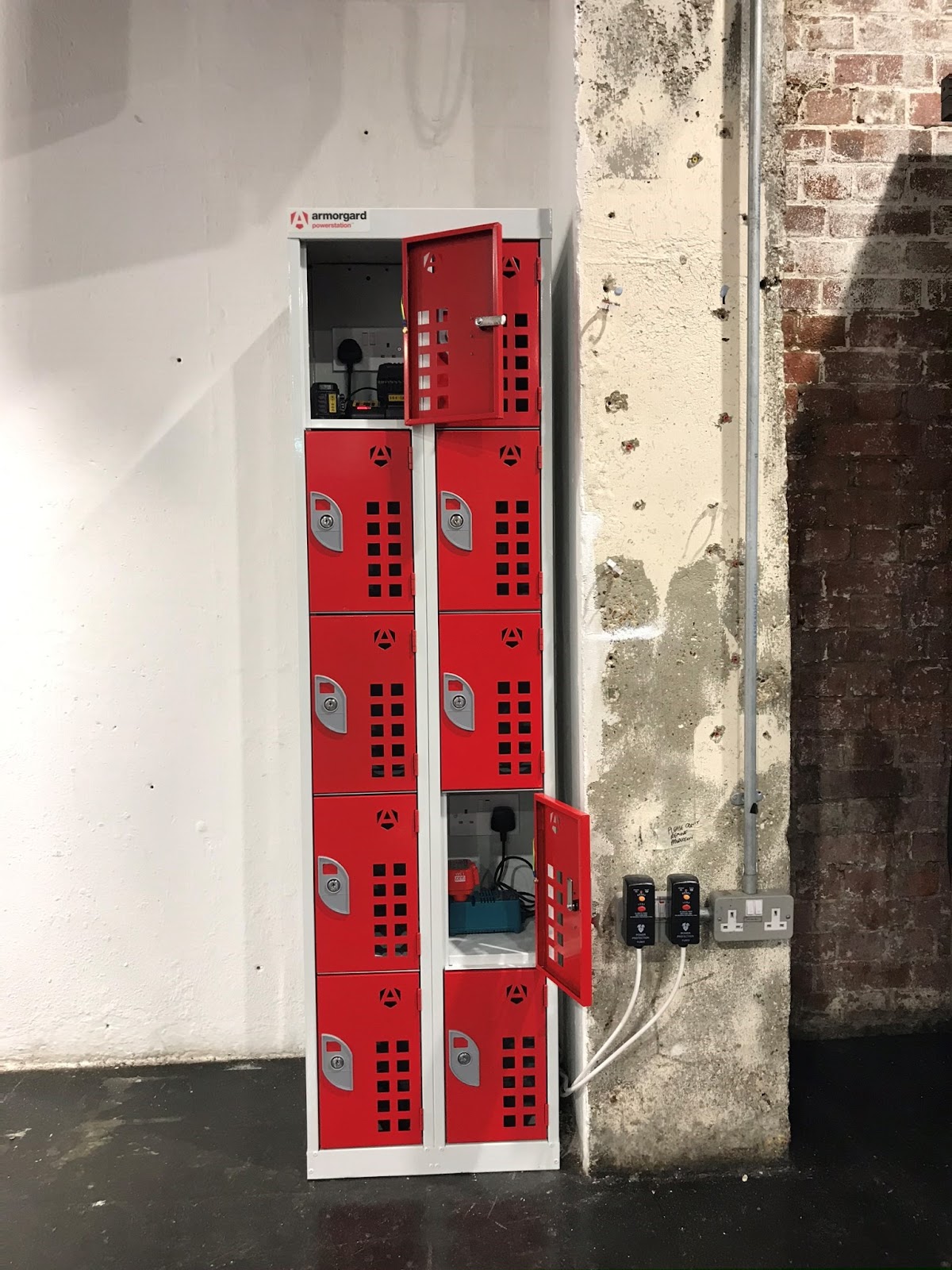 A locker with red doors on a building site, couple of the doors are open and you can see equipment being charged inside the lockers