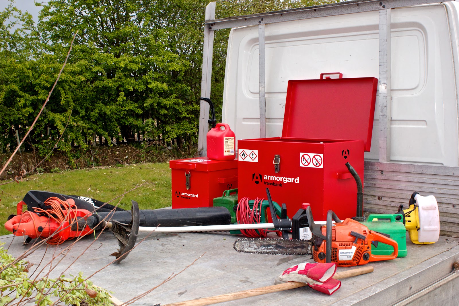 An open transbank on the flat bed of a white van, surrounded by fuel canisters, a chainsaw and a strimmer aswell as some other related equipment. there is also some greenery in the background
