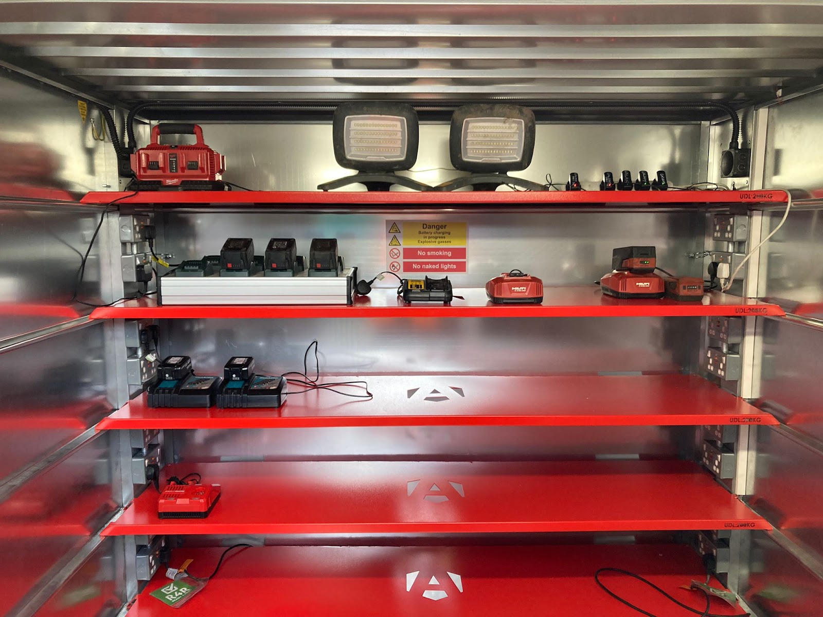 Inside a storage unit with 5 red shelfs and power sockets on both sides of each shelves, there is also equipment being charged on a few of those shelves