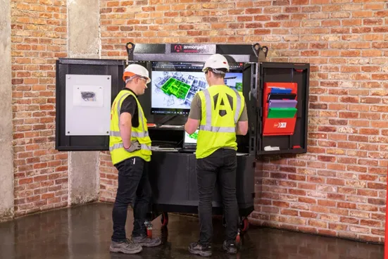 two people wearing hi vis vests and hard hats appear to be discussing a design shown on an open datastation with an exposed brick wall as the background