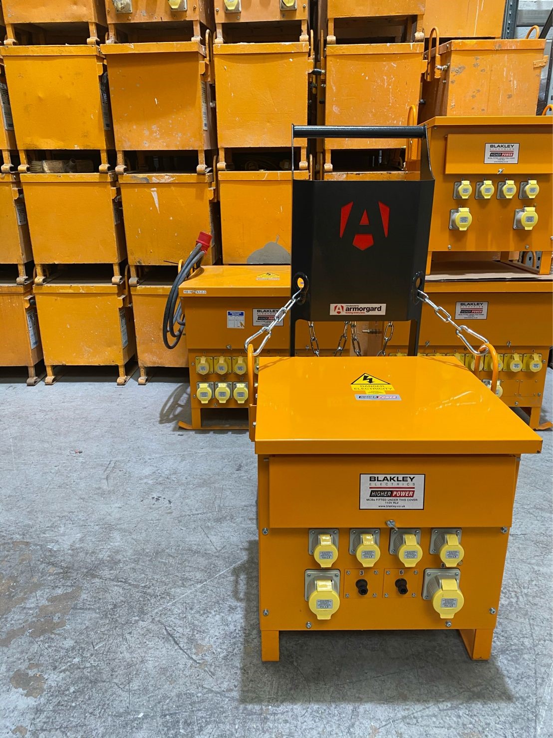 A T-Kart with a site transformer attached to it by chains and in the background there is a big stack of more yellow site transformers