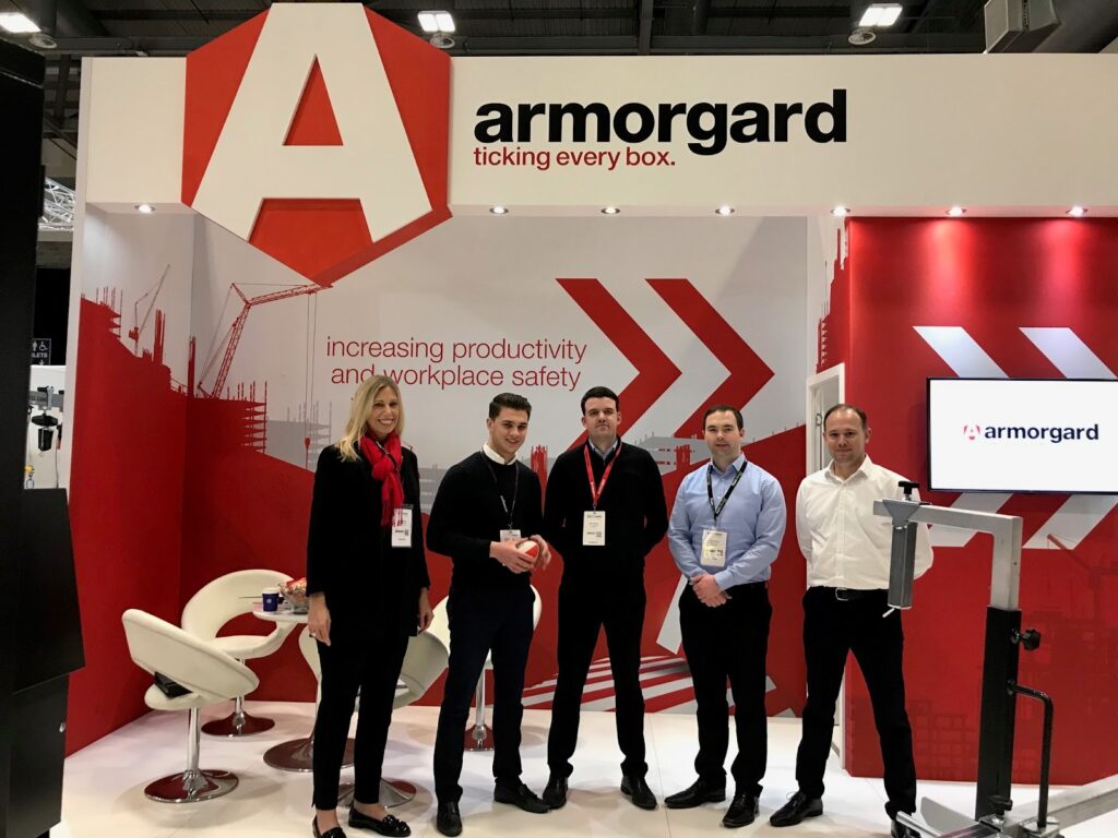 5 Staff members from Armorgard are standing in front of their red and white themed Armorgard stand at the executive hire show.