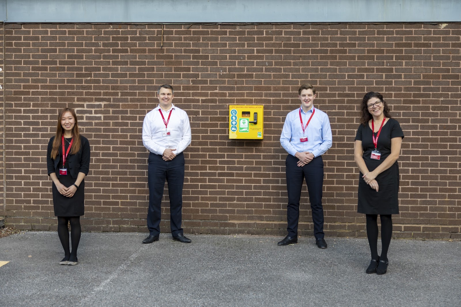4 smiling Armorgard employees wearing red armorgard lanyards standing next to the defibrilator on Armorgard premises