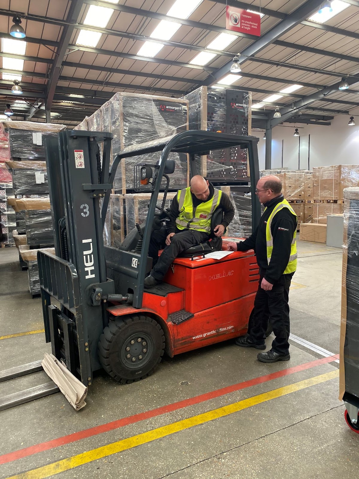 Two people in hi vis vests inside a warehouse surrounded by Armorgard stock, there is one person on in a forklift and another person standing next to it talking and showing something a piece of paper