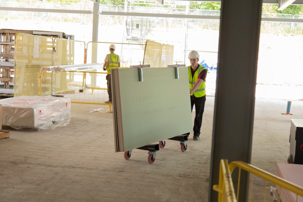 A worker in a hi-vis vest and a white helmet is pushing some large sheet materials on the Armorgard LoadAll trolly