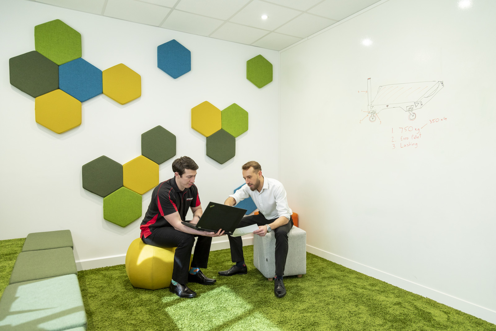 Two people sitting next to each other and one person is holding a laptop and the other is holding paperwork and pointing at the laptop screen.