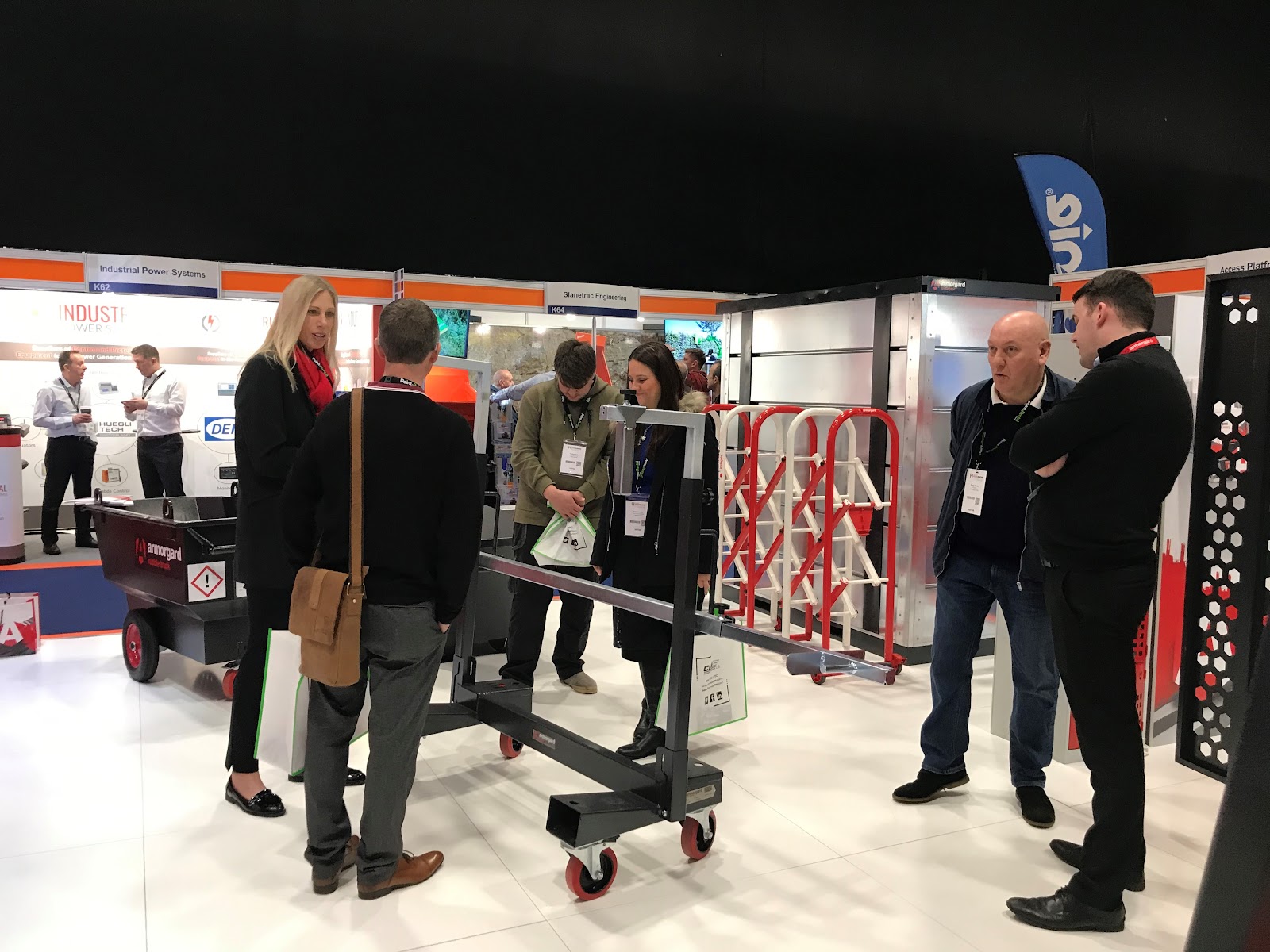 6 people gathering around an Armorgard LoadAll board trolley, which has adjustable clamps on some swivel wheels.