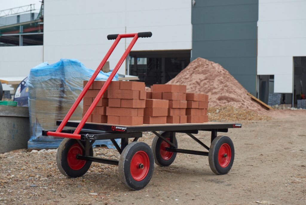 A TurnTable truck in red and black, which has large rubber wheels with a pile of bricks on top of it. There is also a long handle bar to pull the TurnTable Truck.