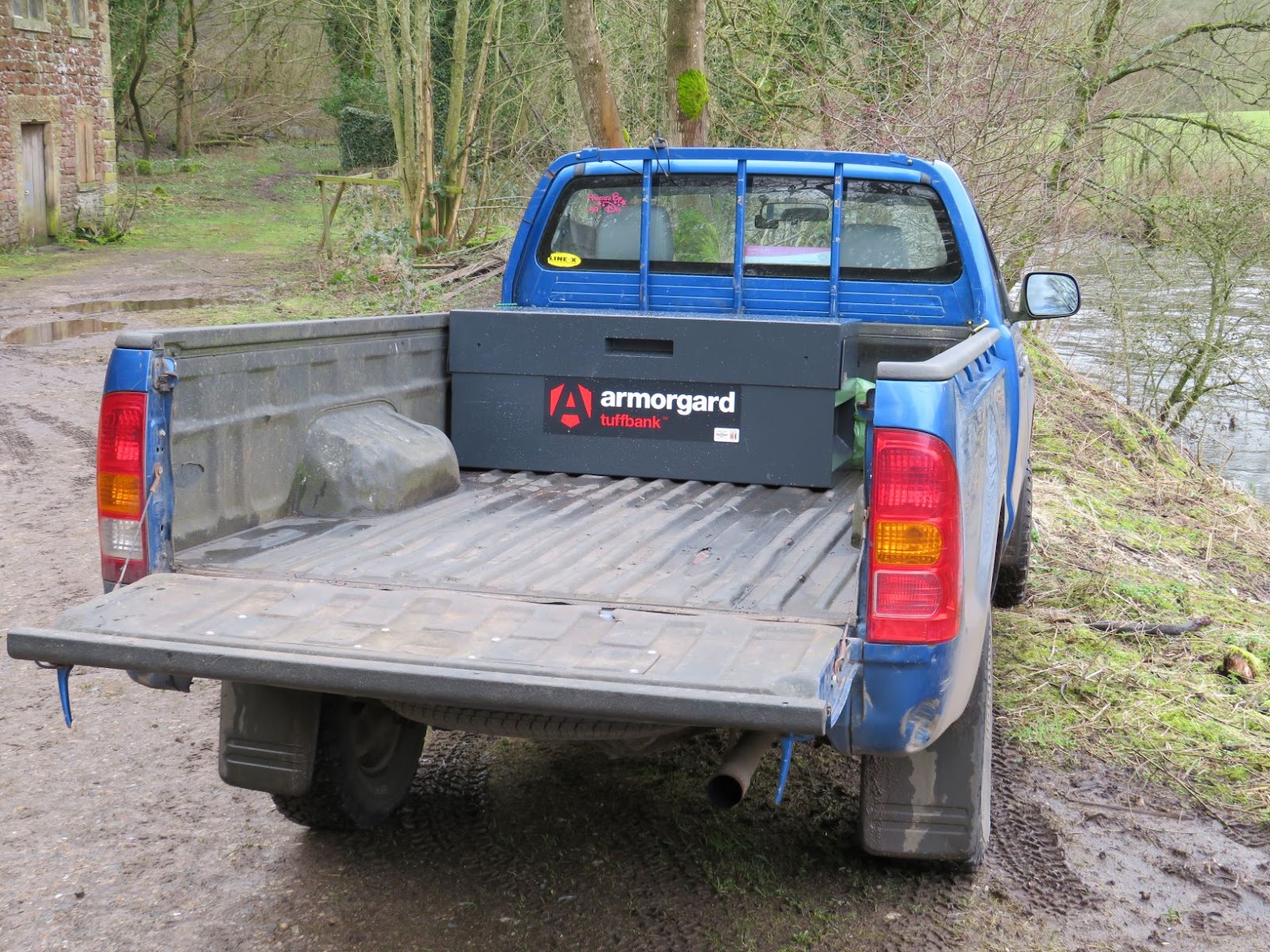 An Armorgard Tuffbank tool chest on the back of a blue pick up truck which is on a slightly muddy road next to a river
