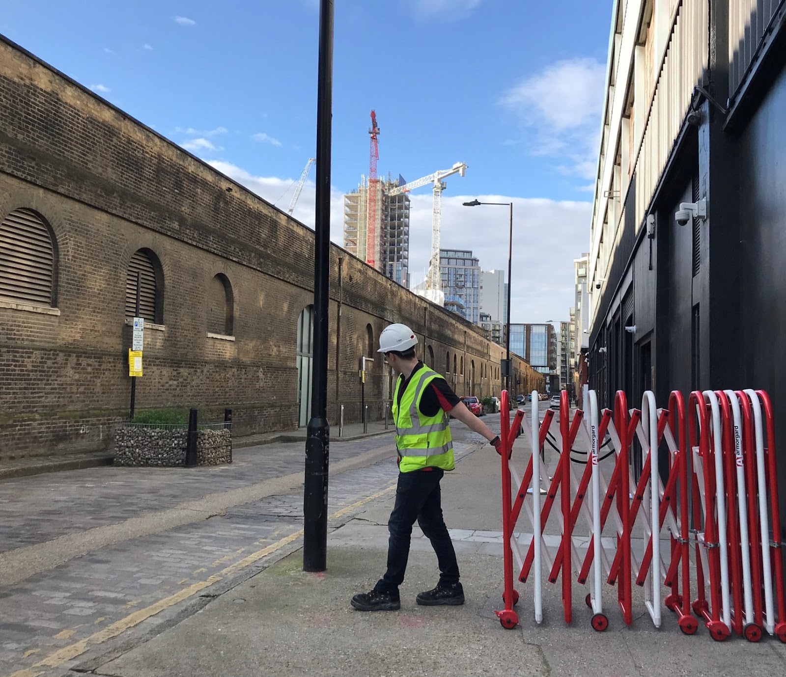 Construction worker in a high-visibility vest and hard hat moving a red and white Armorgard expandable barrier along a city street with buildings and cranes in the background