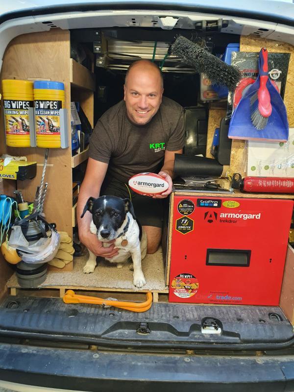 A person kneeling down in the back of a van surrounded by cleaning equipment and an armorgard trekdror, the man is holding a small armorgrad branded rugby ball and there is also a small black and white dog just in front of him