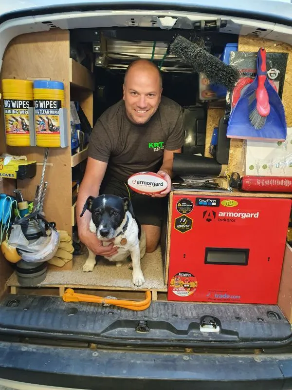 A person kneeling down in the back of a van surrounded by cleaning equipment and an armorgard trekdror, the man is holding a small armorgrad branded rugby ball and there is also a small black and white dog just in front of him