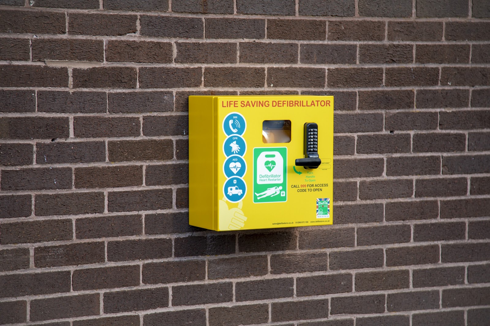 A metal yellow box on a brick wall with a passcode lock on it which contains a defibrillator